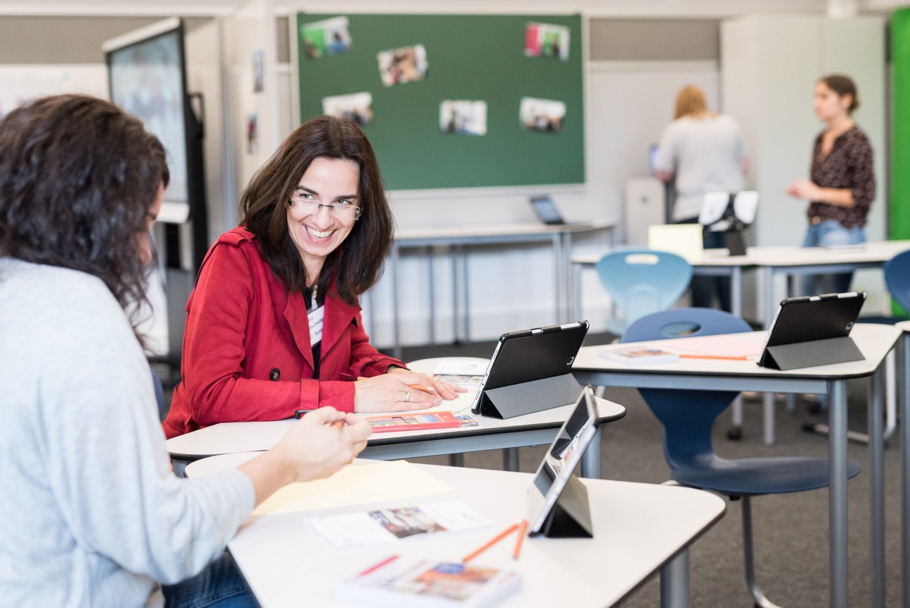 Lehrerin bespricht sich mit einer Kollegin an Tisch mit Tablet und Lernmaterialien im Klassenzimmer.