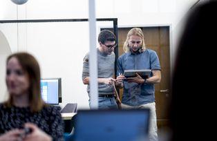 Zwei Männer schauen auf einen Laptop, im Vordergrund diskutieren Menschen