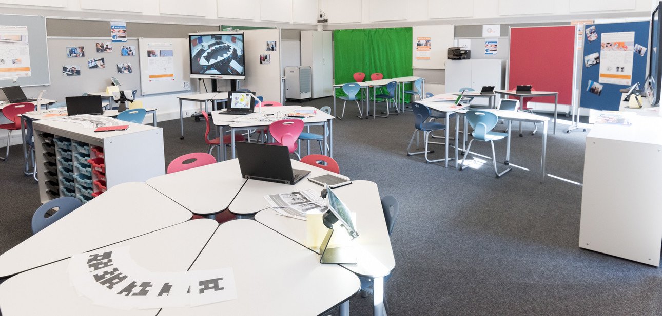 Ein modernes Klassenzimmer mit flexiblen Tischen, bunten St&uuml;hlen, Laptops und interaktiven Whiteboards. Die Ausstattung unterst&uuml;tzt digitales Lernen und kollaborative Arbeitsmethoden.