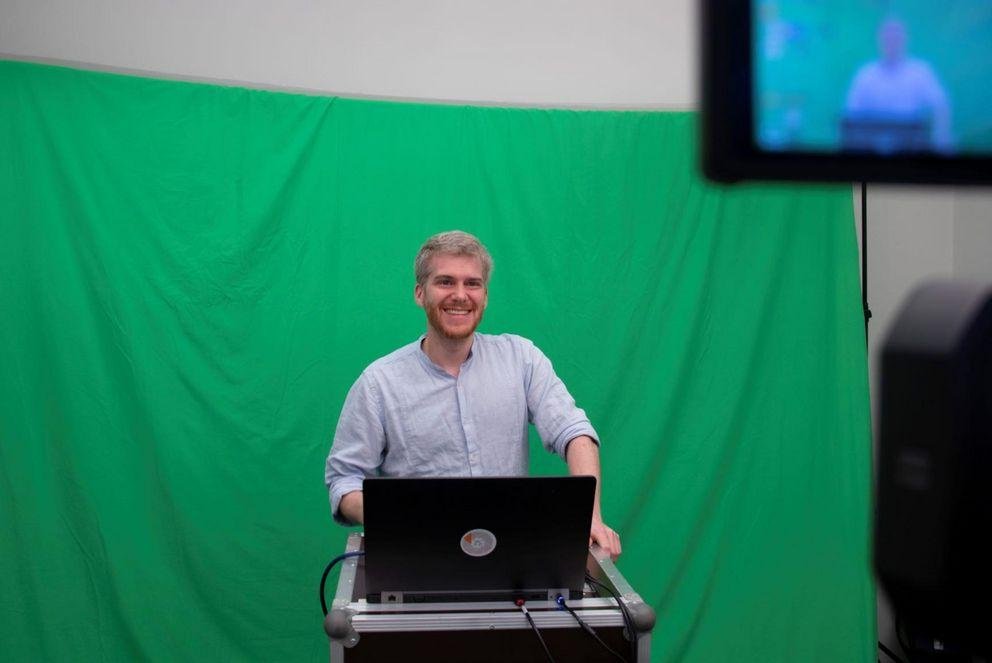 Dozent steht vor einem Greenscreen und lächelt am Rednerpult in die Kamera. Die Kamera ist am rechten oberen Bildrand zu erkennen, wie sie die Szene aufnimmt.
