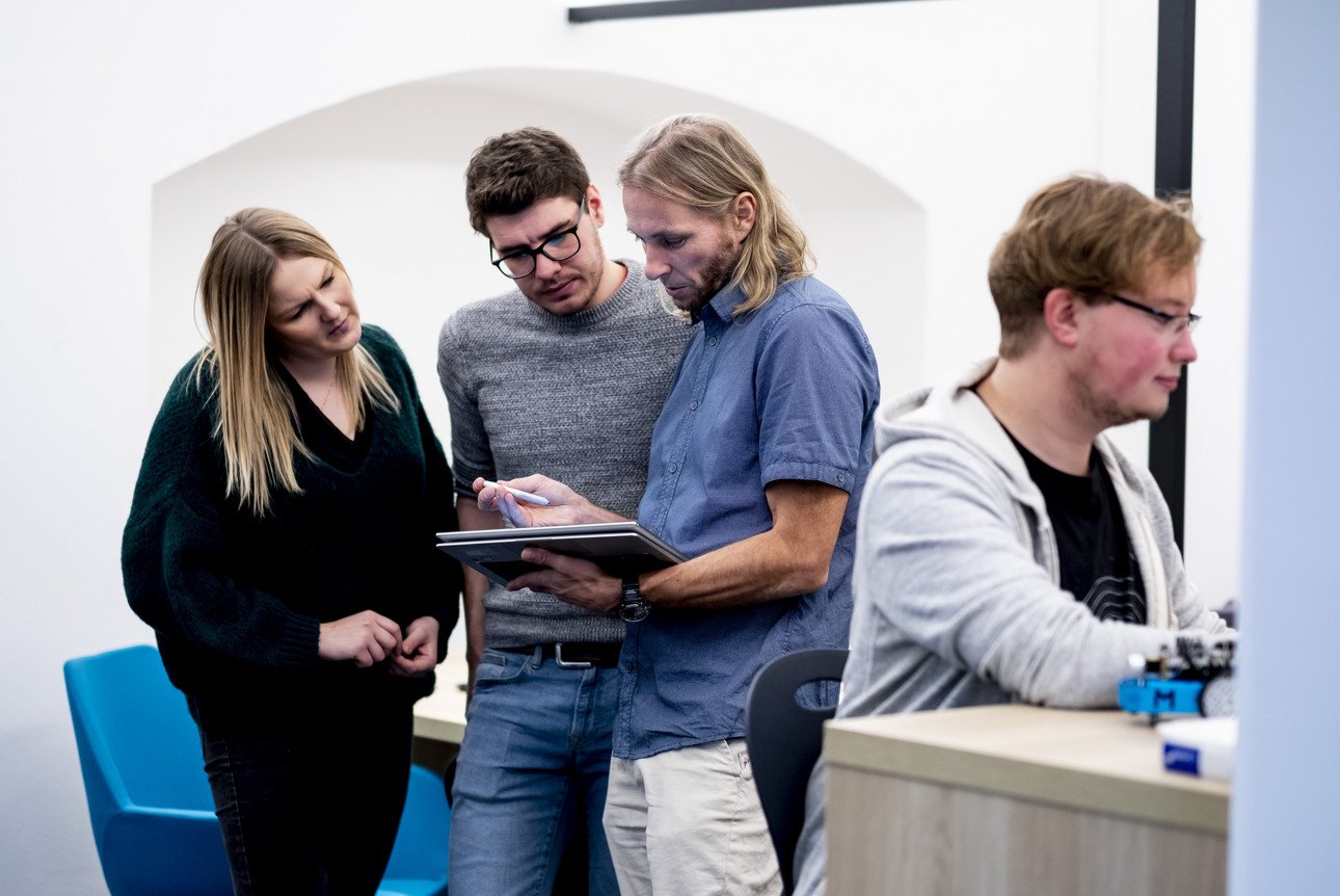 Vier Lehrkr&auml;fte in einem modernen B&uuml;roarbeitsumfeld, die intensiv an einem Tablet zusammenarbeiten und strategisch &uuml;ber ein Projekt diskutieren.
